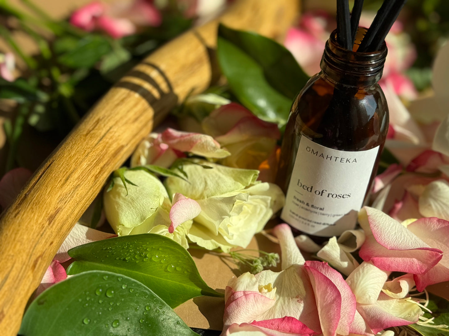 Bed of Roses de la Omahteka – parfum de cameră cu bețișoare, într-un cadru cu frunze picurate de rouă, plante verzi și petale de trandafiri.