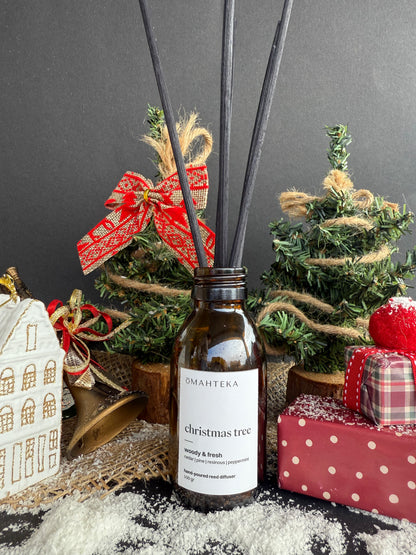 Difuzorul de cameră Christmas Tree de la Omahteka, fotografiat cu bețișoare negre și decoruri de Crăciun cu brad și globuri.