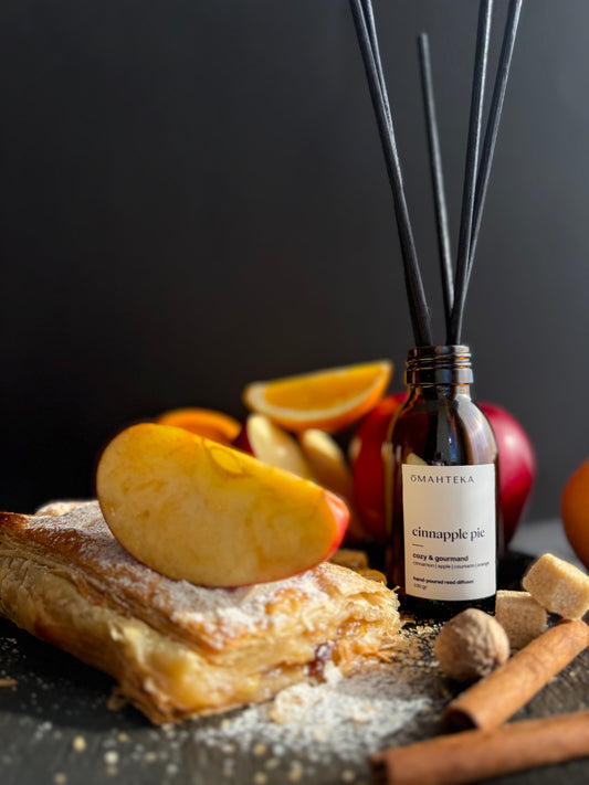 Difuzorul de cameră Cinnapple Pie de la Omahteka, prezentat cu bețișoare negre din ratan și decor cu plăcintă de mere și scorțișoară.