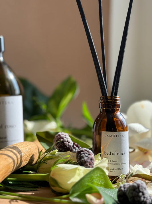 Parfum de cameră Bed of Roses de la Omahteka – difuzor cu bețișoare din ratan, în sticlă maro cu protecție UV, plasat într-un decor floral cu trandafiri și frunze verzi.