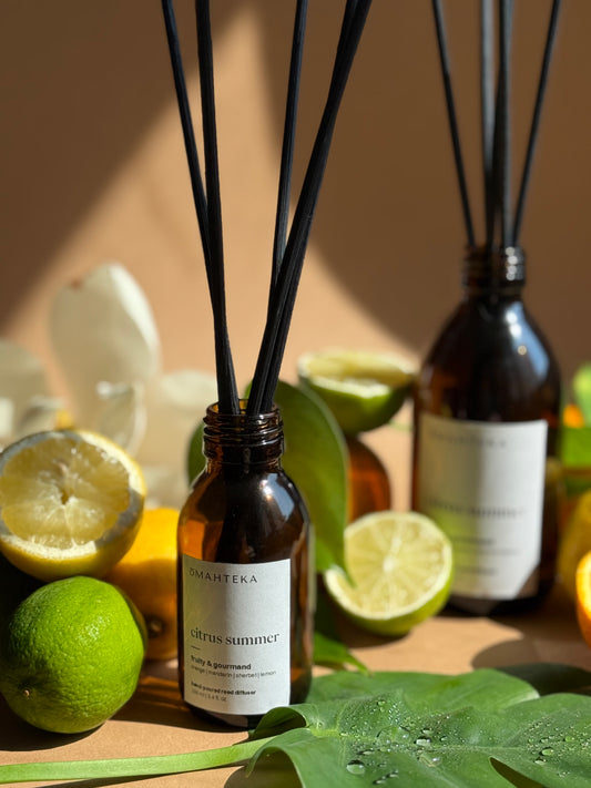 Parfum de cameră cu bețișoare Omahteka – Citrus Summer, într-un flacon de sticlă maro cu protecție UV, înconjurat de lime, portocale și frunze verzi.